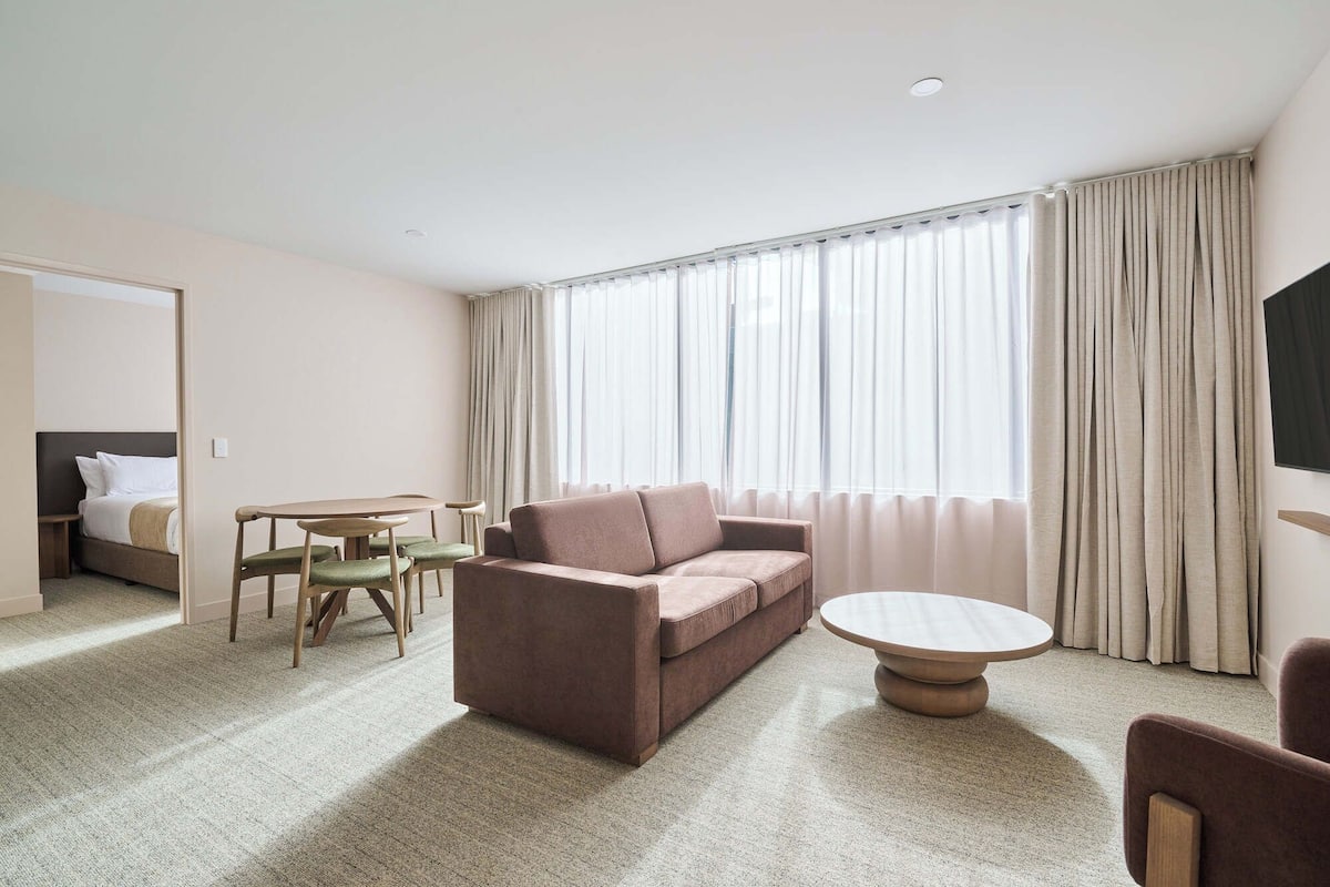 A hotel room with a sofa, a round coffee table, a small dining table with chairs, and a bed with a headboard.