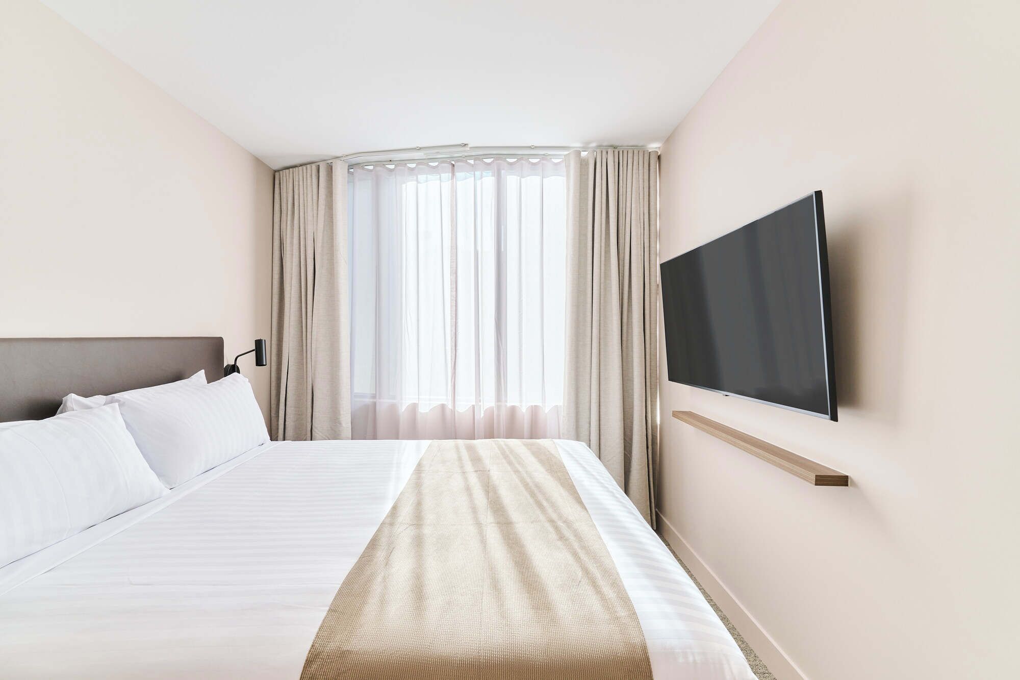 Ein Hotelzimmer mit einem Bett, einem an der Wand montierten Fernseher und einem Fenster mit Vorhängen.