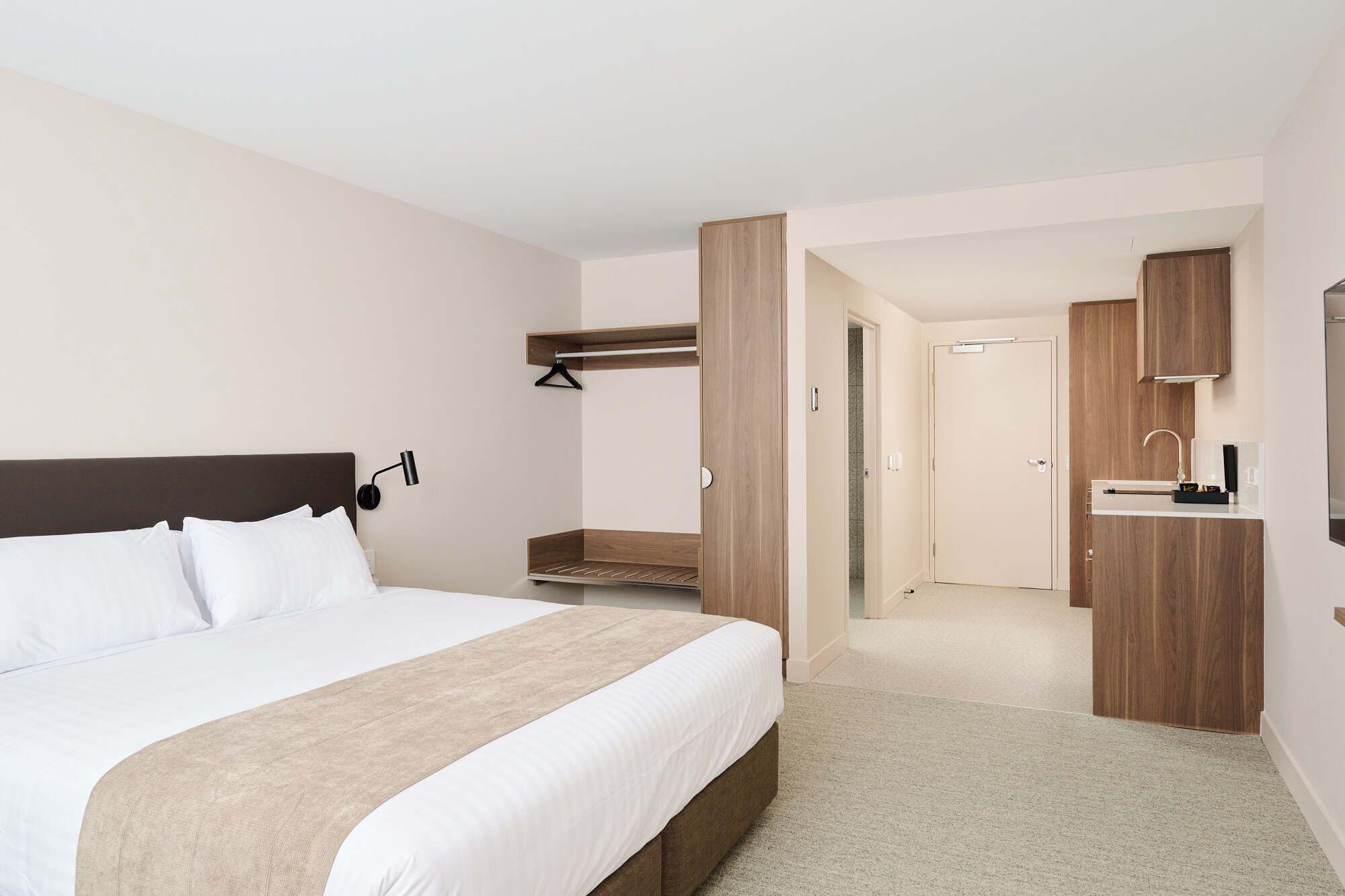 Une chambre d’hôtel avec un grand lit, une table de chevet, une étagère murale, une salle de bain avec un lavabo et un miroir, et une porte donnant sur une autre pièce.