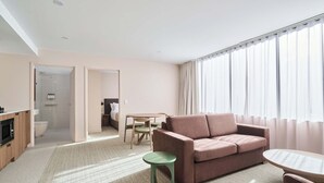 A modern hotel room with a sofa, a small round table, a green stool, a shower area, and a bathroom. - Abode Phillip (Phillip)
