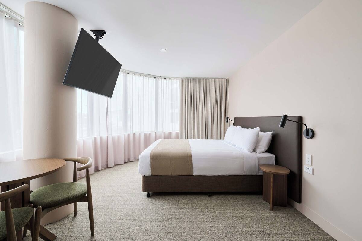 A hotel room with a bed, a desk, a chair, a TV mounted on the wall, and a window with curtains.