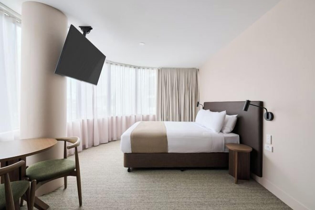 A hotel room with a bed, a desk, a chair, a TV mounted on the wall, and a window with curtains.