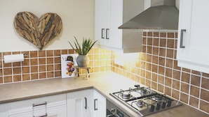 A kitchen with white cabinets, a gas stove, a heart-shaped wall decoration, and a potted plant on a small stool. - Immaculate 1-bed Cottage in Moreton in Marsh (Moreton-in-Marsh)
