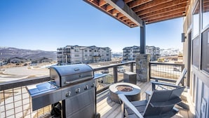 Ein Balkon mit Grill und Gartenmöbeln, mit Blick auf ein Wohngebiet mit mehreren Gebäuden und einer Bergkulisse.