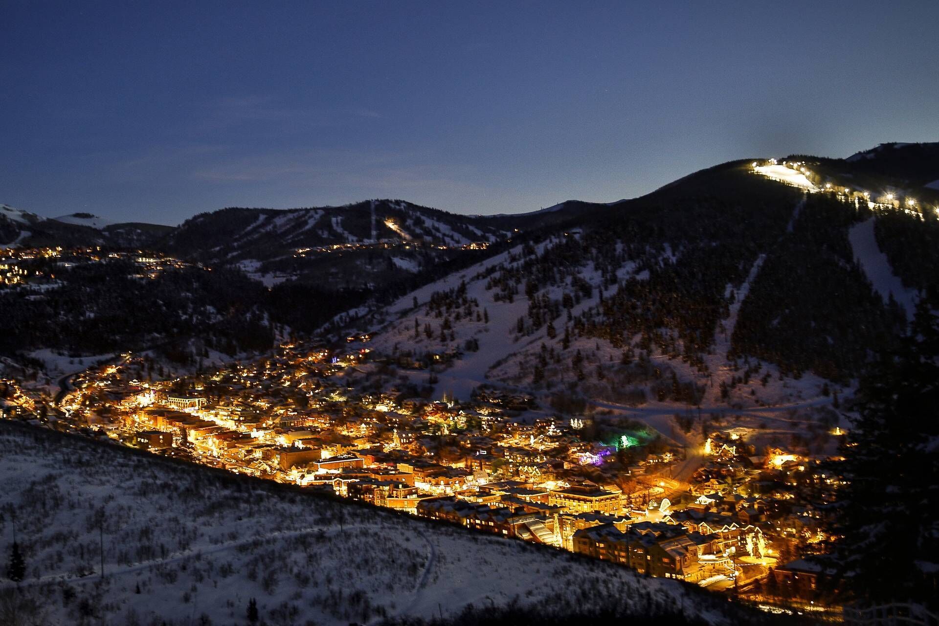 A snow-covered mountain town at night with illuminated buildings and a view of the surrounding mountains.