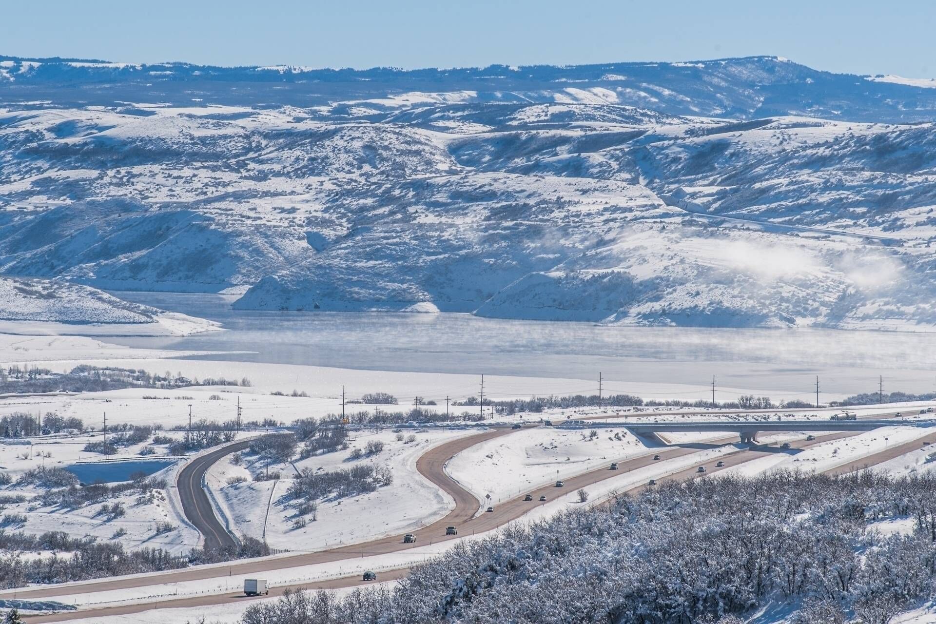 A snow-covered landscape with a highway curving through it, a bridge, and distant mountains.