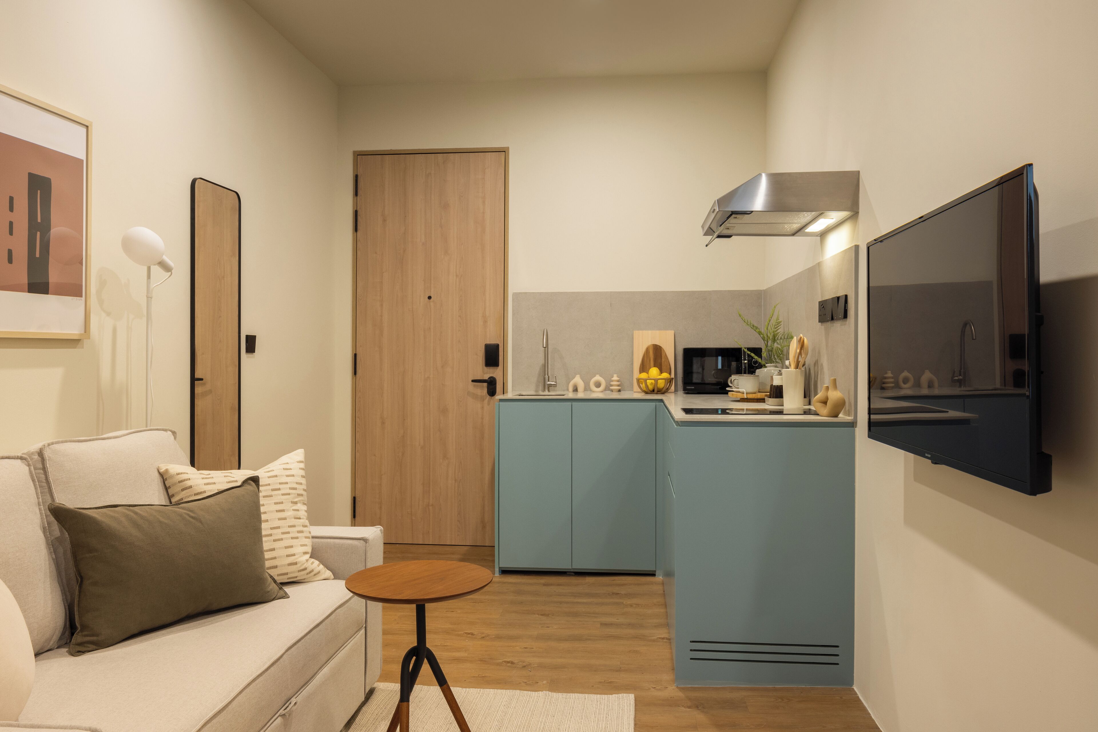 Un espace de vie compact comprenant un coin cuisine, un canapé avec des coussins, une petite table ronde et un téléviseur fixé au mur.