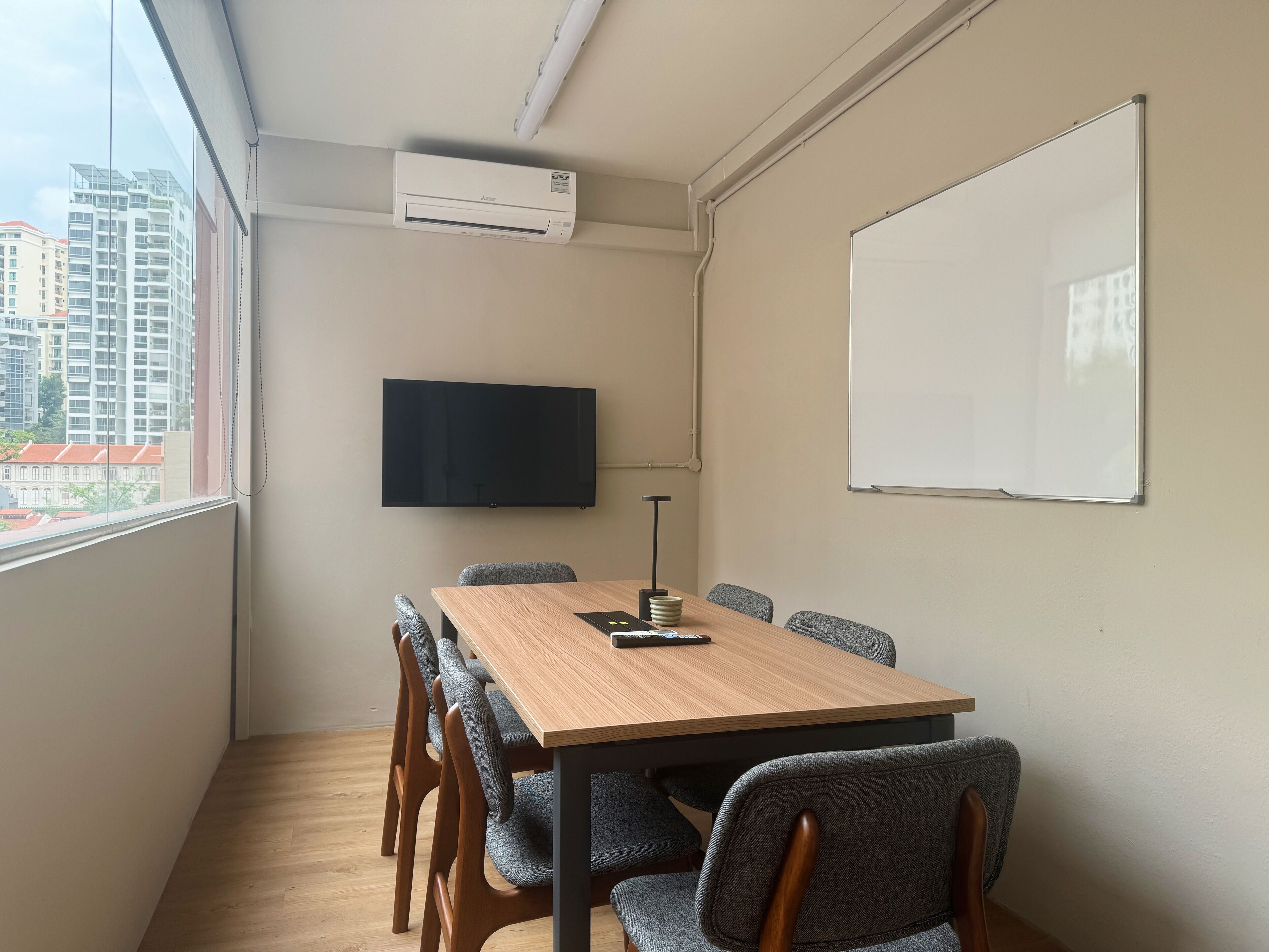A conference room with a wooden table, grey upholstered chairs, a mounted flat-screen TV, a whiteboard, and an air conditioning unit.