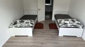 Una habitación pequeña y estrecha con dos camas individuales, una puerta que da a un pasillo y una ventana sobre la puerta.