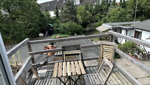 Una terraza de madera con una mesa y sillas, rodeada de follaje y un edificio al fondo.
