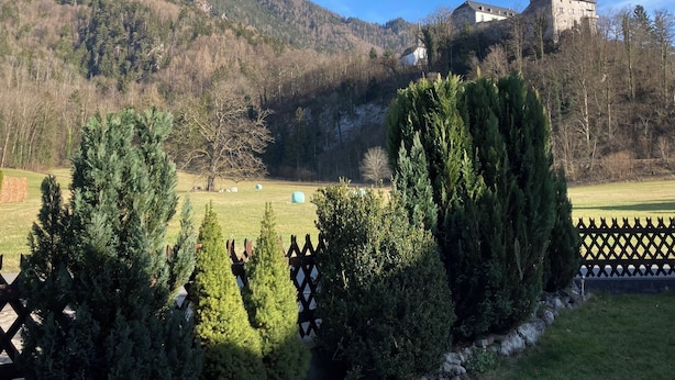 Paisaje montañoso con una edificación de estilo castillo en una colina, rodeado de árboles y un área cercada con fardos de heno.