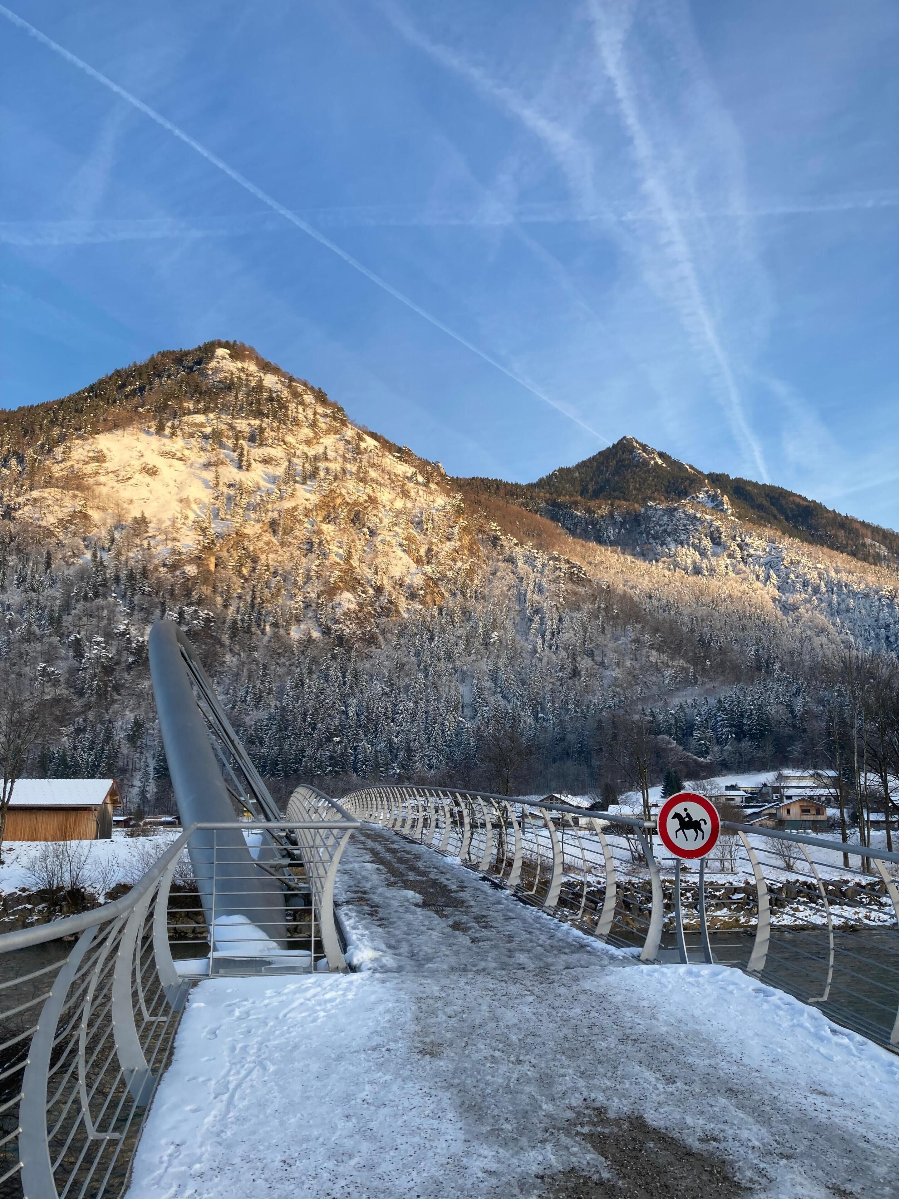 Een sneeuwbedekte brug met een metalen hekwerk, een bord met daarop 'verboden voor paarden' en een bergachtige achtergrond.