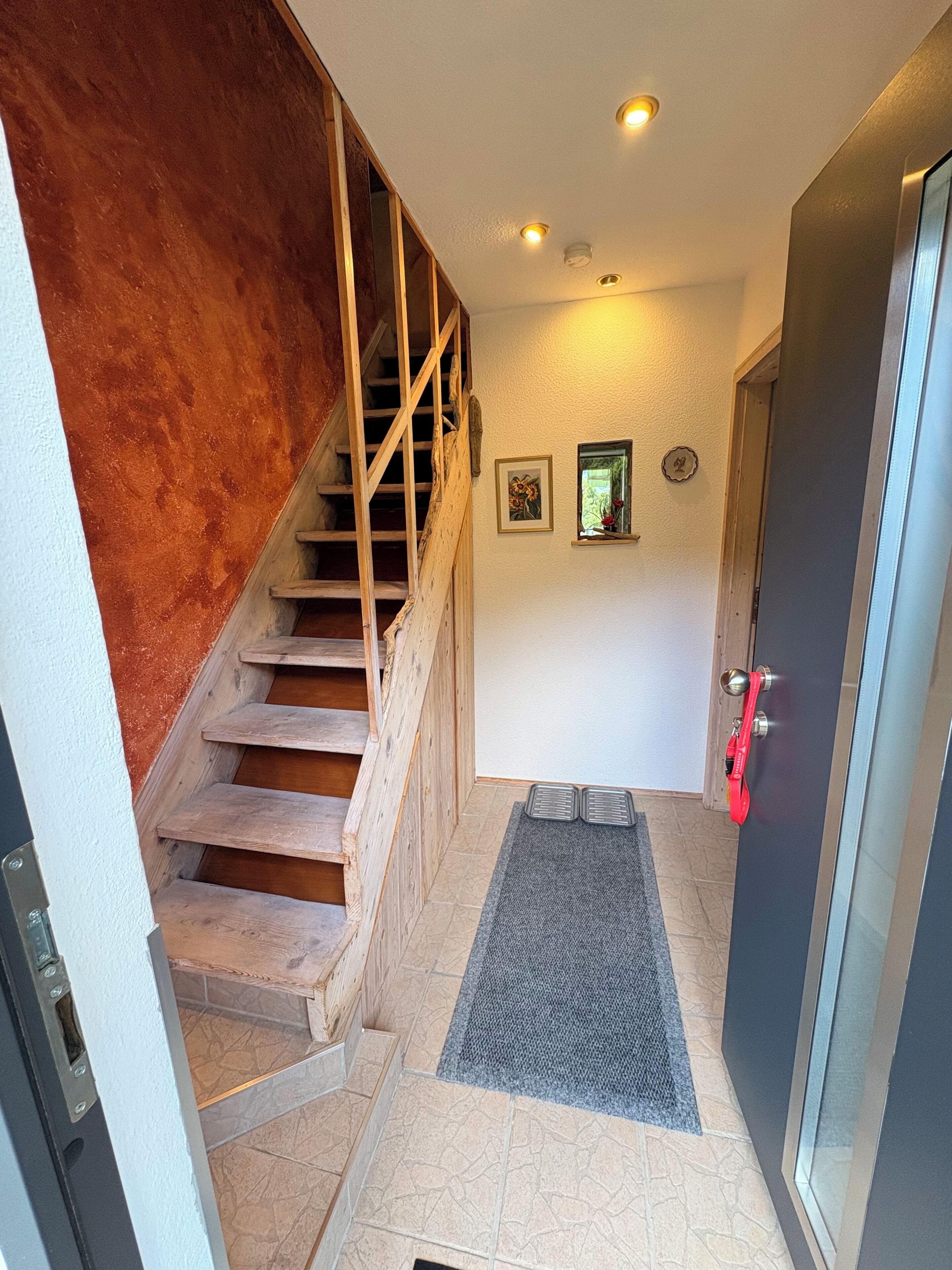 Eine Treppe mit Holzstufen führt zu einer oberen Etage, eine Tür mit rotem Griff und ein kleines Fenster mit einer Pflanze.