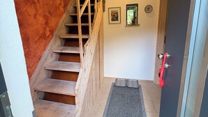 Una escalera de madera que sube a un piso superior, una puerta con picaporte rojo y una ventana pequeña con una planta.