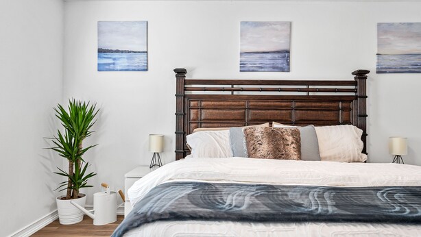 A bedroom with a wooden headboard, a large bed with patterned bedding, a potted plant, bedside lamps, and three wall-mounted paintings. - Beautiful and cozy apartment in Bonita w/Garden  (Bonita Springs)
