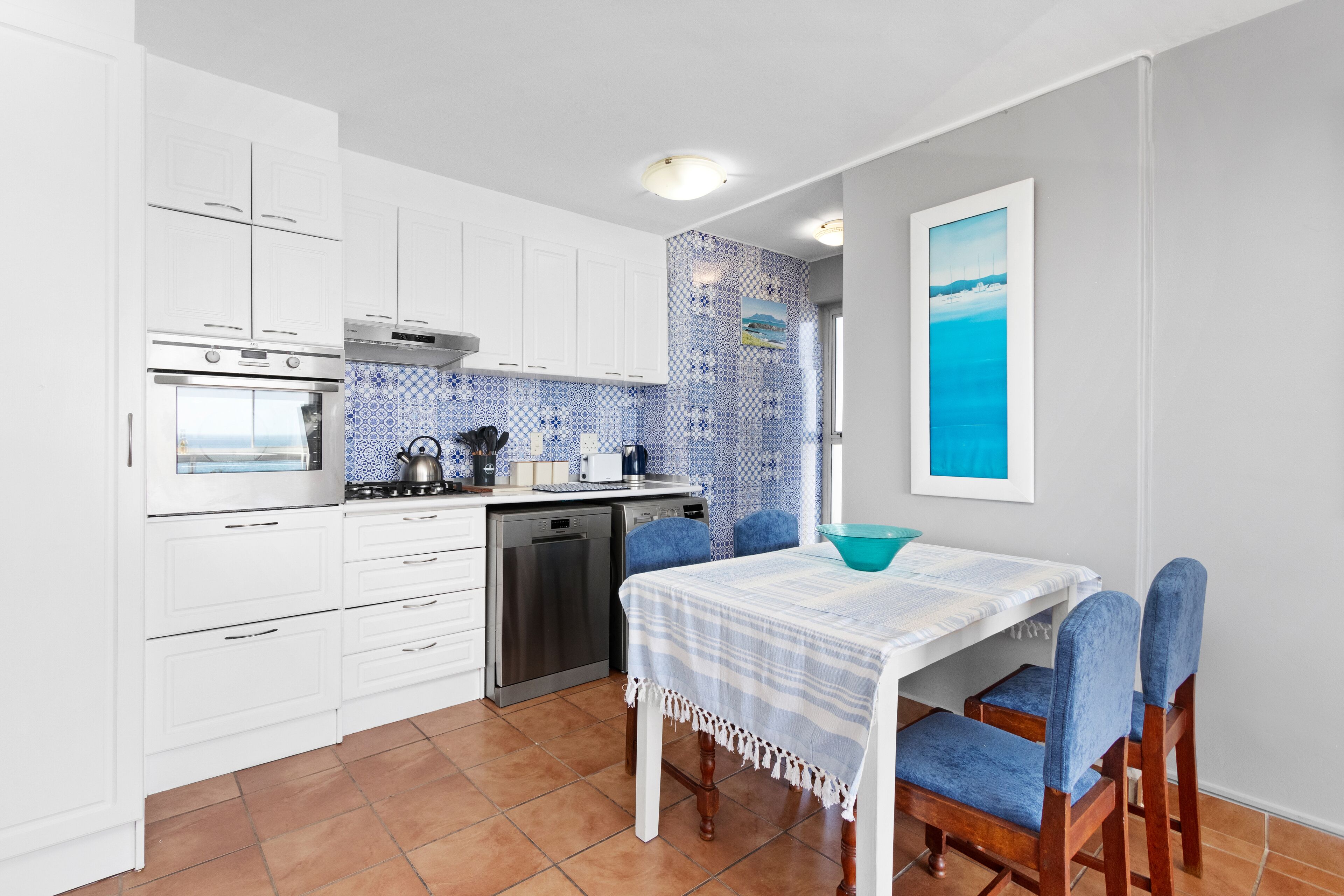 A kitchen with white cabinets, a stove, and a dishwasher. There is a table with a blue bowl and chairs.