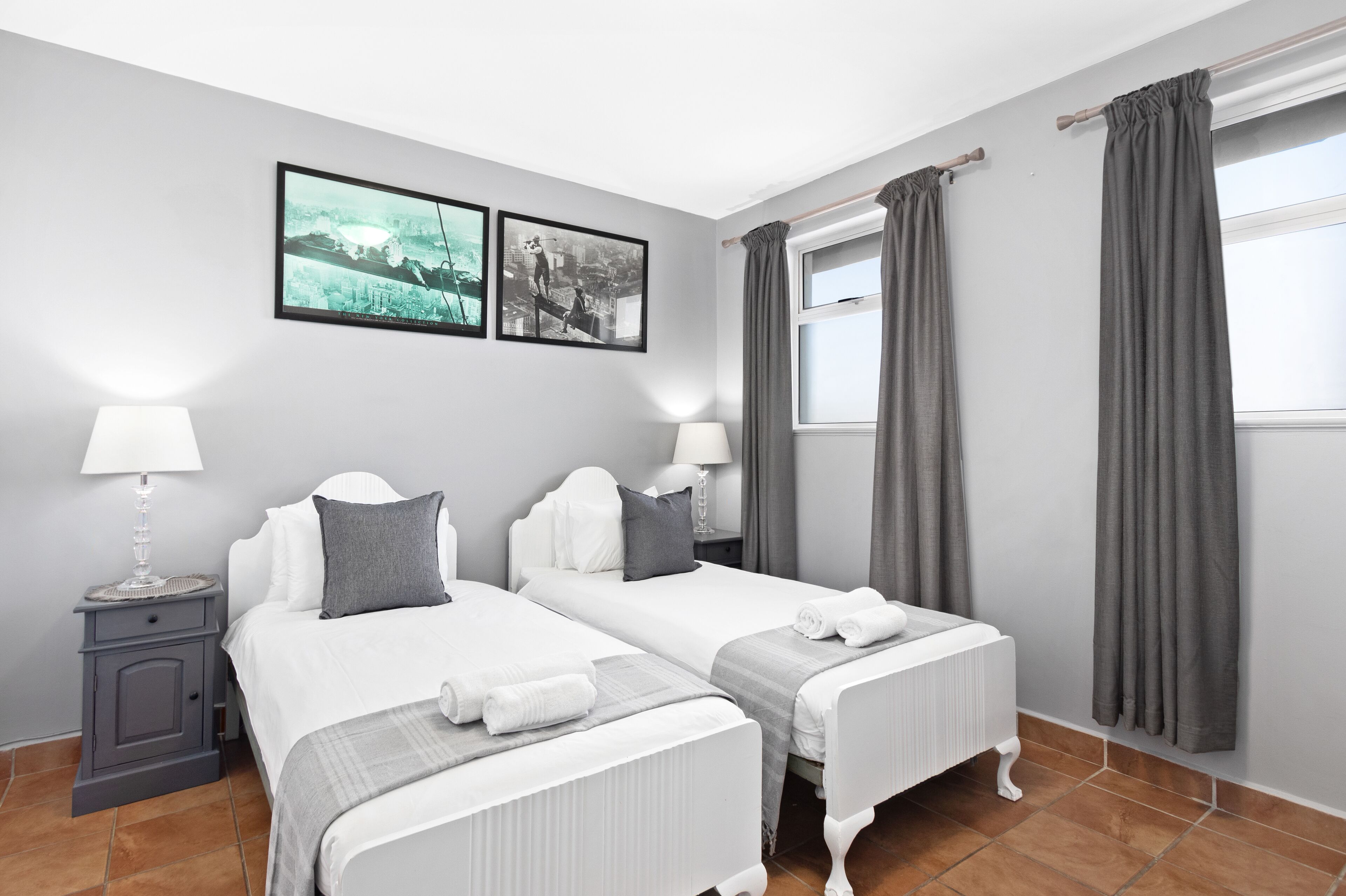 Quarto de hotel com duas camas, cortinas cinzentas, uma obra de arte com tema urbano e piso ladrilhado.