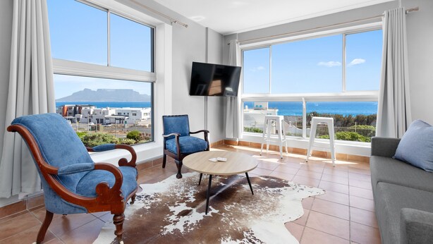 Un salon moderne avec une grande télévision, deux fauteuils bleus, une table basse ronde et une vue sur la mer.