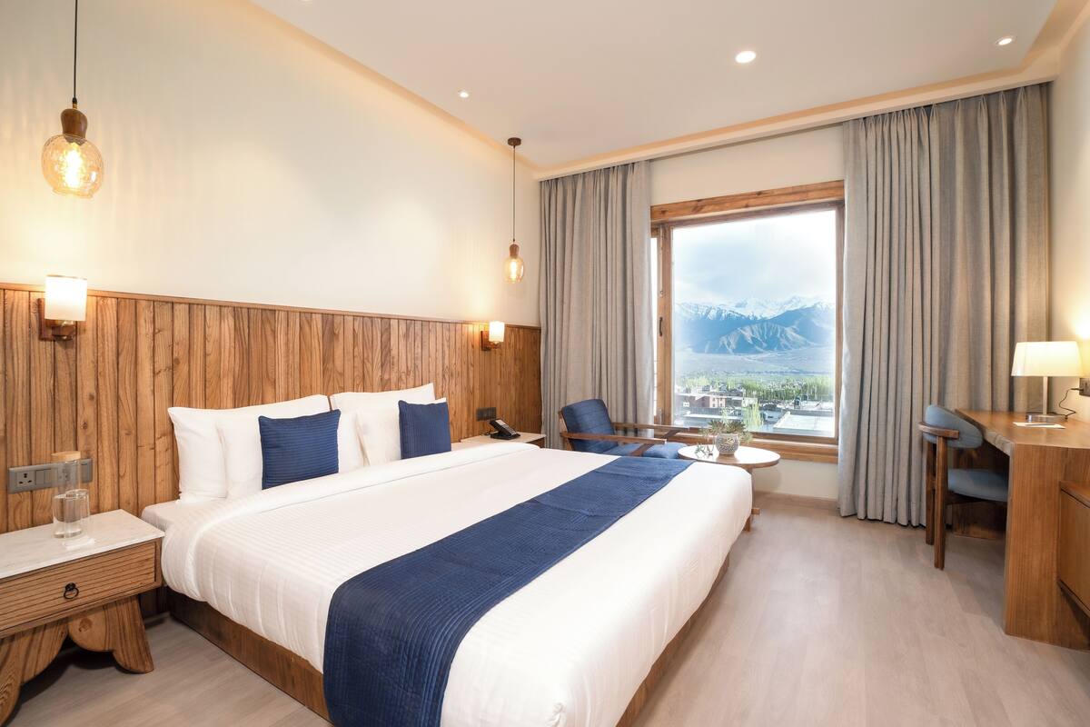 A hotel room with a large bed, a desk, a chair, a small table, and a view of mountains through the window.