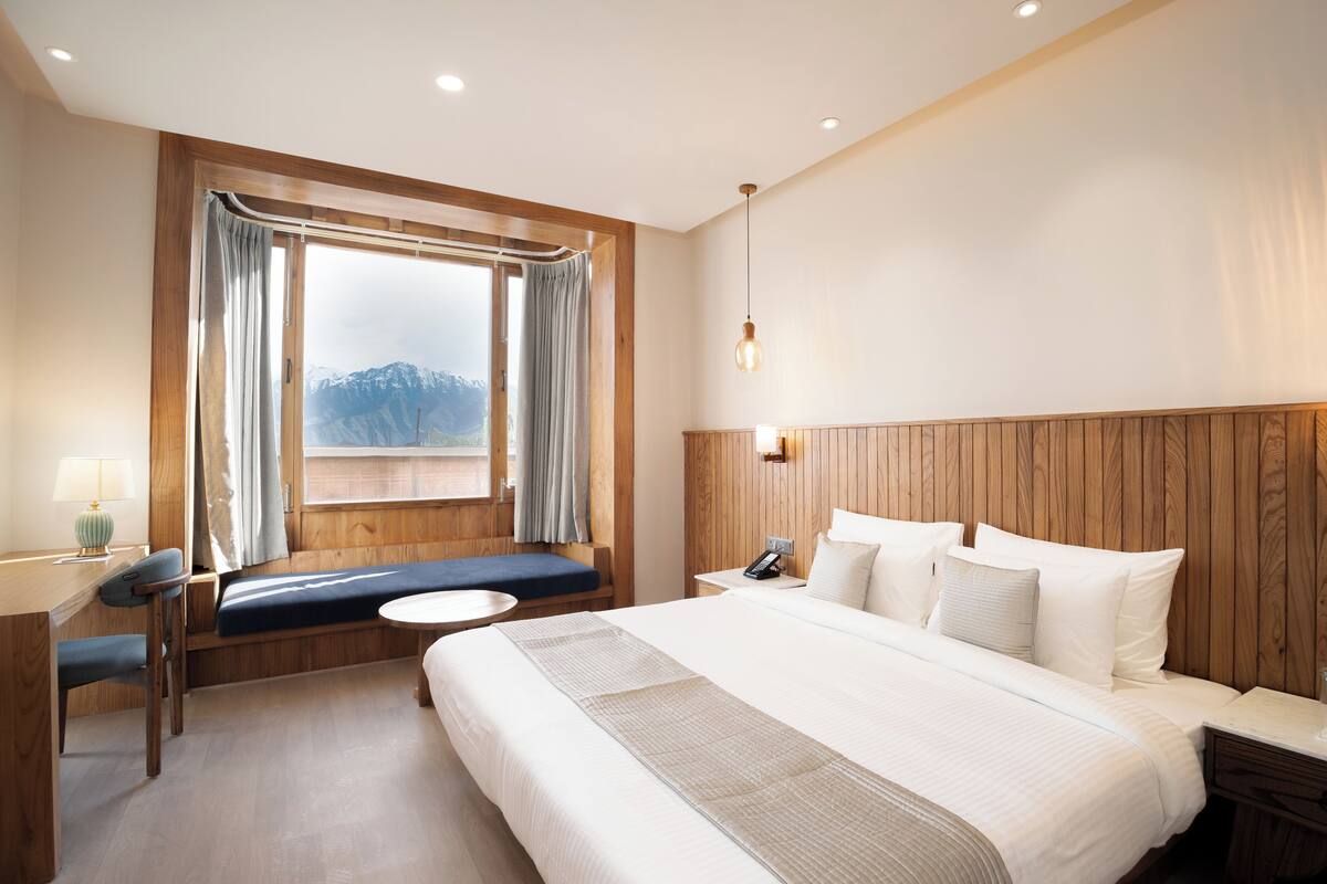 A hotel room with a large bed, a wooden headboard, a window with curtains, a desk with a lamp, and a view of mountains.