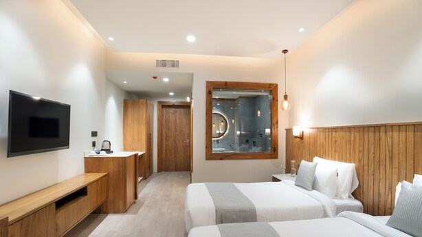 Habitación de hotel moderna con dos camas, televisor, cabecero de madera y baño visible a través de una puerta de cristal.