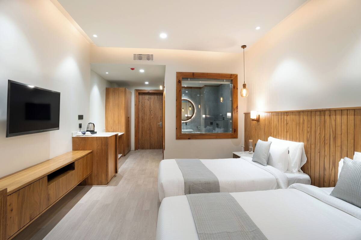 A modern hotel room with two beds, a TV, a wooden headboard, and a bathroom visible through a glass door.