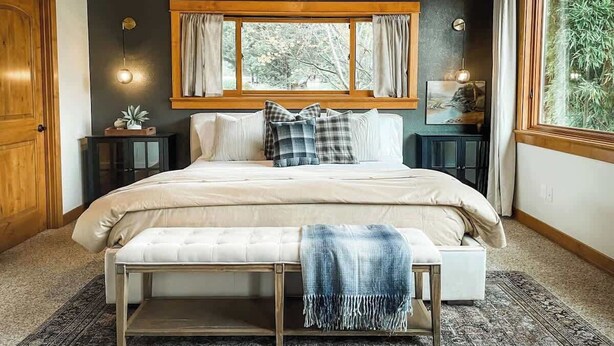 Quarto com uma cama grande, um banquinho, uma mesa de cabeceira, um relógio de parede e uma janela com cortinas.