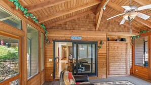 Interior de uma cabana de madeira, com um ventilador de teto, uma placa com a inscrição 'God Bless' e uma porta de vidro que dá para outro cômodo.