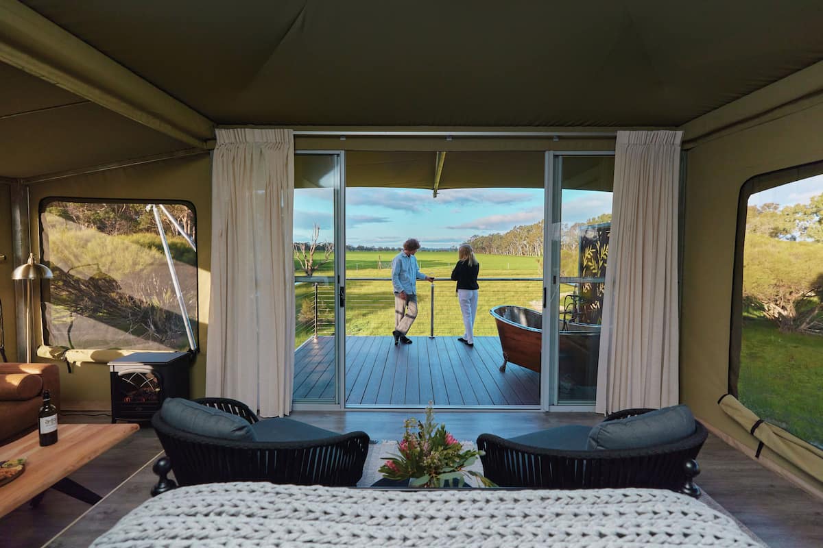 Een kamer met een grote schuifglasdeur die uitkomt op een terras. Binnen zijn een bed, twee stoelen, een kleine tafel met een fles en een decoratieve plant.