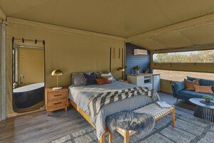 Un dormitorio con cama, mesita de noche, lámpara, banco y vistas al exterior.