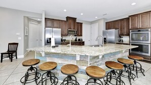 A modern kitchen with granite countertops, stainless steel appliances, wooden bar stools, and a large island with a sink. - Disney-Area Home w/ Themed & Media Room, Pool Table & Waterpark - 240CL (Kissimmee)
