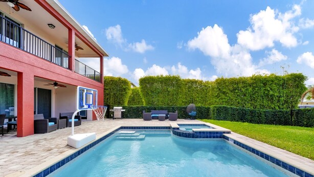 A swimming pool with a red building in the background, a covered patio area with seating, and a well-maintained hedge. - Disney-Area Home w/ Themed & Media Room, Pool Table & Waterpark - 240CL (Kissimmee)