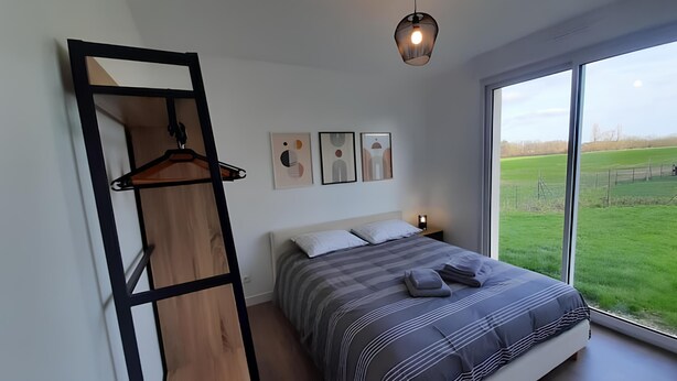 Een slaapkamer met een groot raam, een bed met gestreepte beddengoed, een houten garderobe en abstracte wanddecoratie.