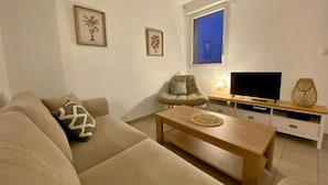 Een woonkamer met een beige bank, een houten salontafel, een tv op een houten dressoir, en een raam met uitzicht op de lucht.