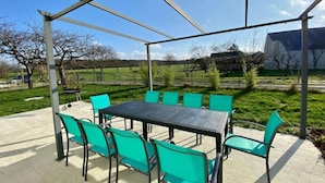 Een overdekt terras met een tafel en stoelen, omgeven door een gazon en bomen.