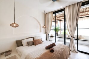 A modern bedroom with a large bed, two hanging pendant lights, and a view of the outdoors through a large window with curtains. - Villa Coconut (Canggu)