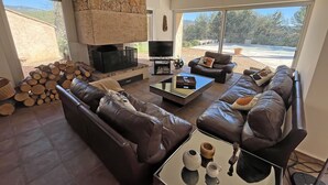 A modern living room with a stone fireplace, leather sofas, a flat-screen TV, and a large window with a view of the outdoors. - Villa San Michele - 70's Experience (Salernes)