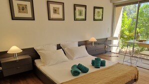 A modern bedroom with a large bed, two bedside tables, a window with a view of greenery, and framed artwork on the wall. - Villa San Michele - 70's Experience (Salernes)