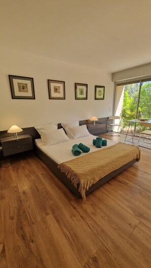 A modern bedroom with a large bed, two bedside tables, a window with a view of greenery, and framed artwork on the wall.