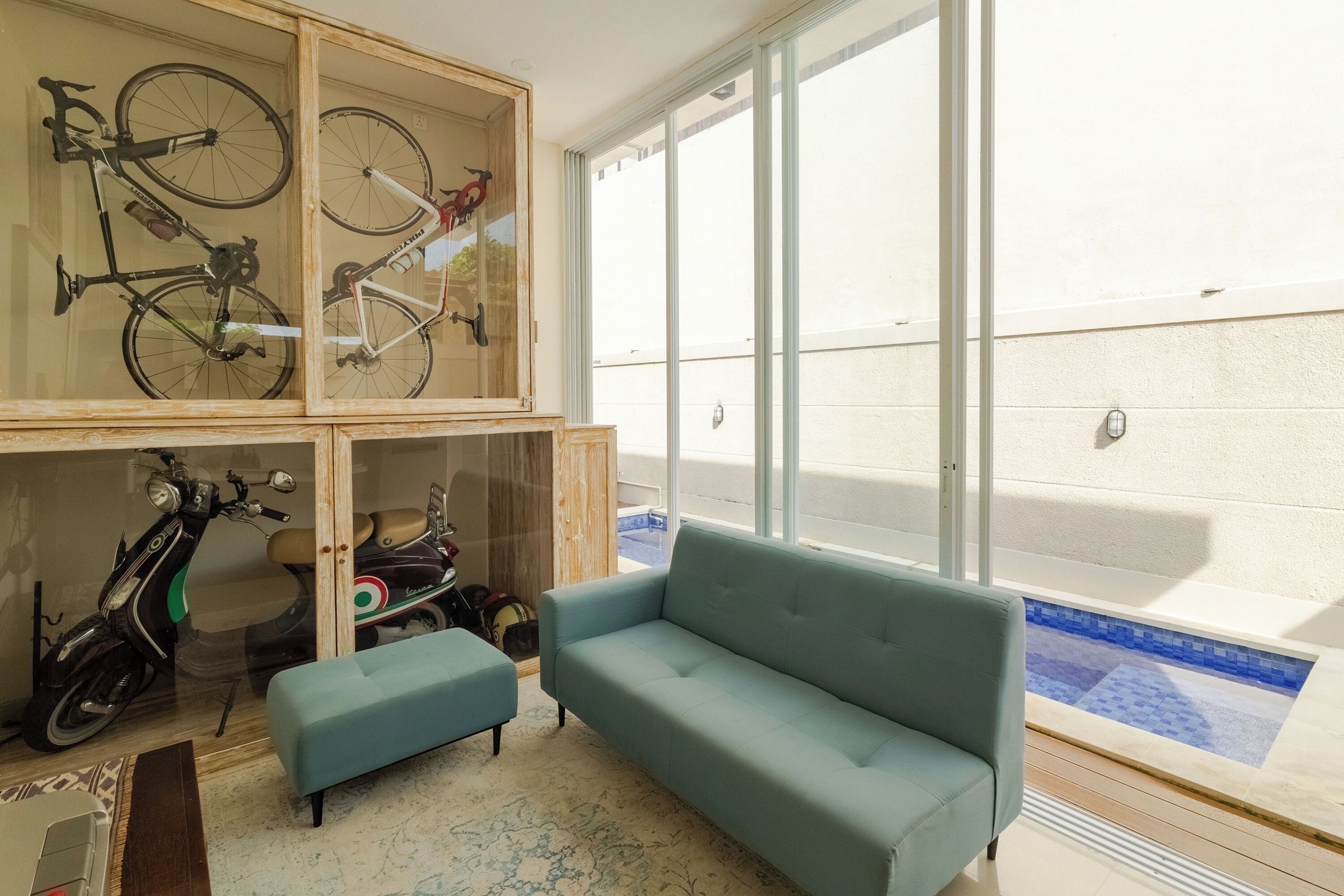 A room with a blue sofa, a wooden cabinet with bicycles and scooters, and a view of a swimming pool through large windows.