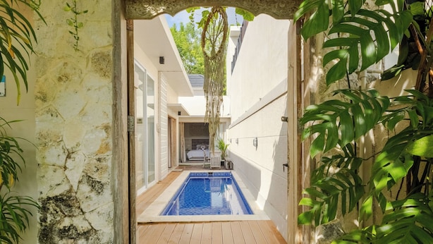 A narrow pathway leading to a rectangular pool with a wooden deck, surrounded by walls with a textured finish and tropical plants. - Rustic Pandawa Villa (Kutuh)