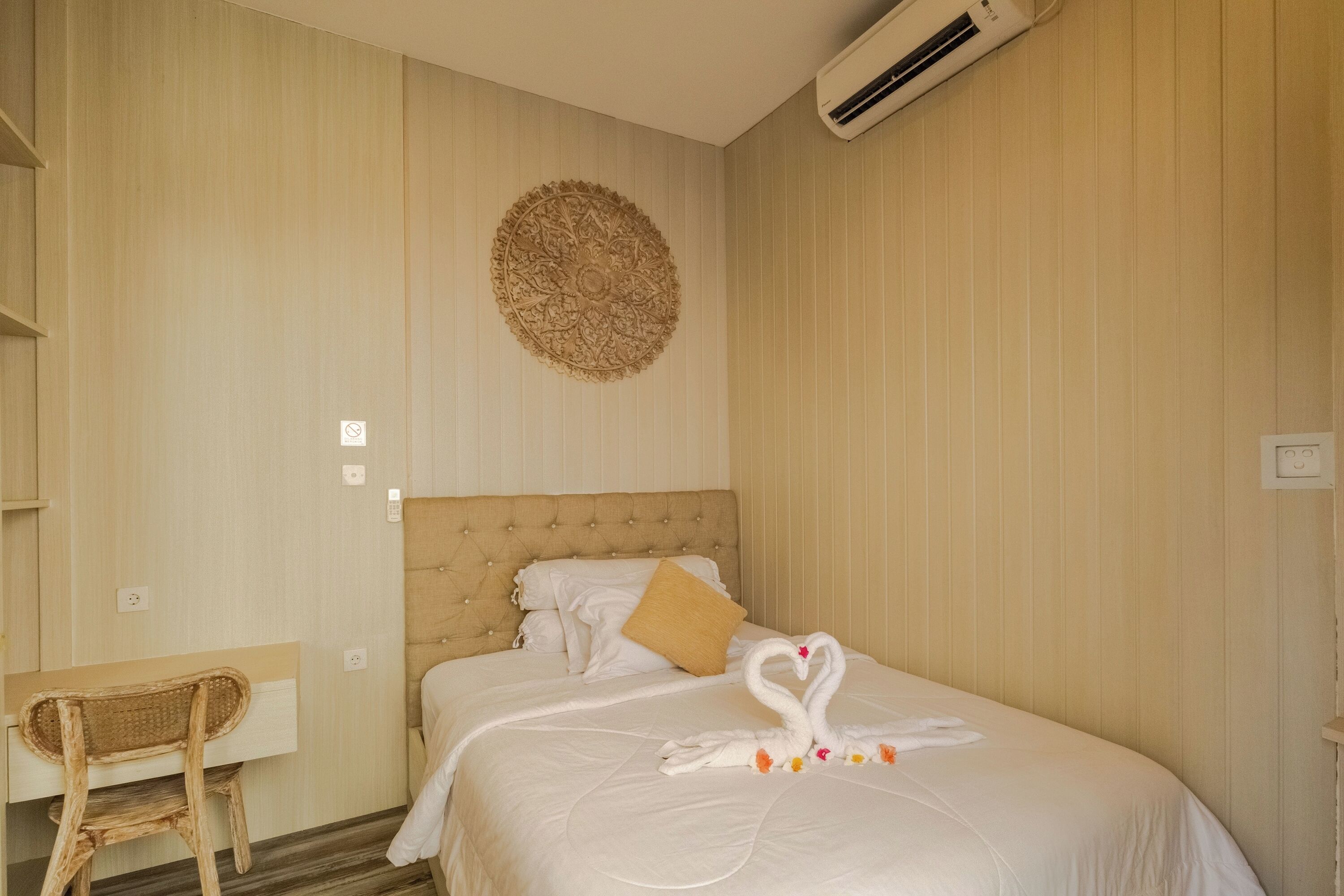 A hotel room with a bed, a wall-mounted air conditioner, a decorative wall piece, a wooden chair, and a bedside table.