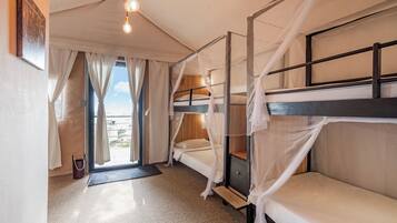A room with two bunk beds, a wooden headboard, a small bedside table, a curtain, and a view of the outdoors.