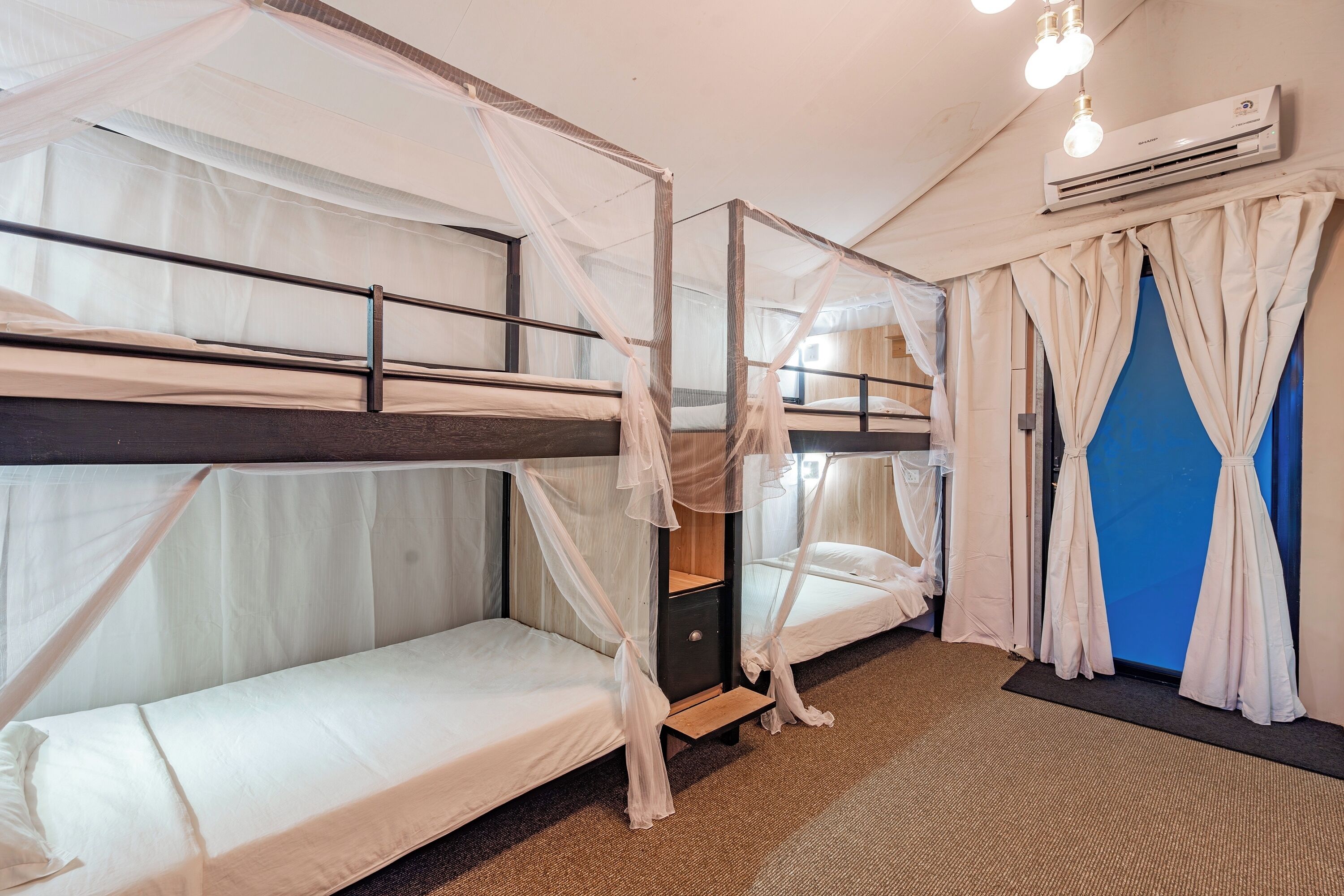 A room with bunk beds, a small wooden nightstand, white curtains, and an air conditioning unit.