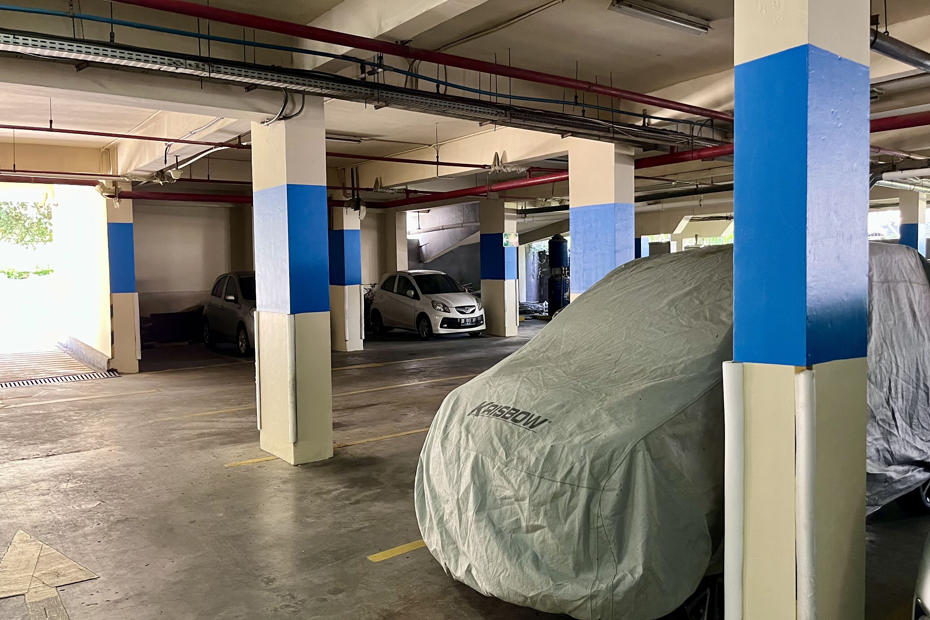 A parking garage with concrete pillars, some covered vehicles, and a car in the background.