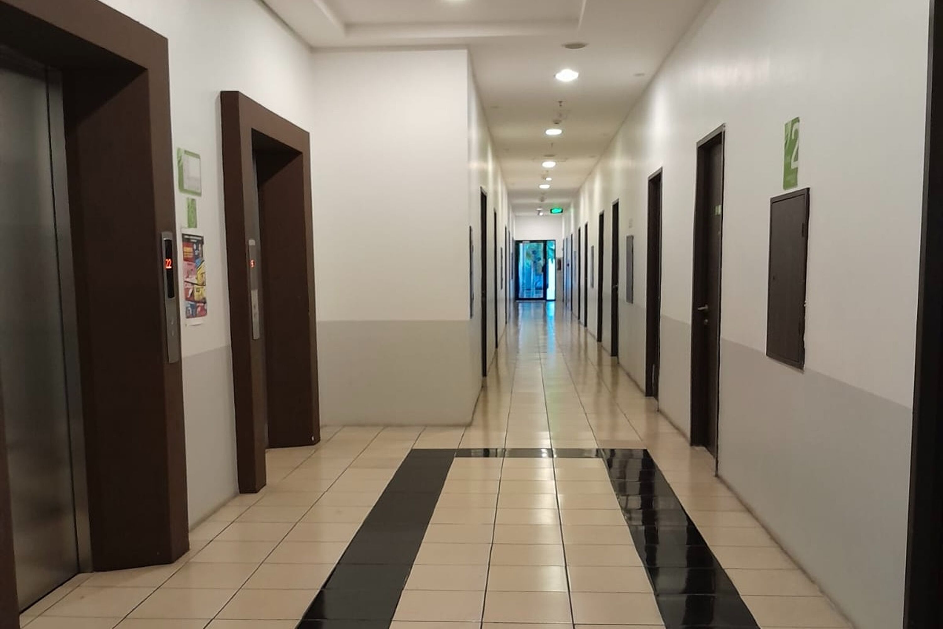 A long, narrow hallway with a black and white tiled floor, multiple doors, and a visible elevator on the left.