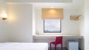 Ein Hotelzimmer mit einem Bett, einem Schreibtisch mit Stuhl, einem kleinen Kühlschrank, einem Fenster mit Blick und einer Wandleuchte.