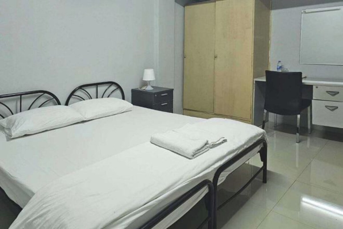 A single-bed room with a metal frame, a bedside table with a lamp, a wardrobe, and a desk with a chair.