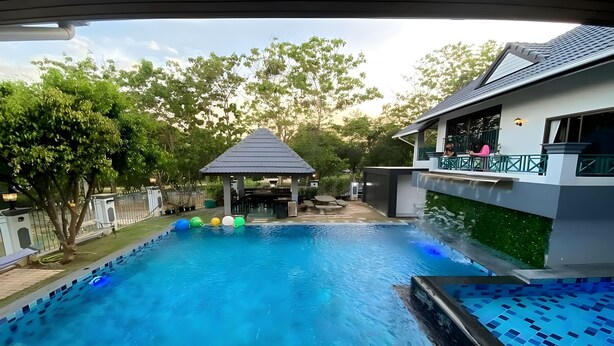 A swimming pool with a gazebo in the background, surrounded by trees and a house with a balcony. - Siriniran Homestay (Sattahip)