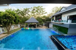 A swimming pool with a gazebo in the background, surrounded by trees and a house with a balcony. - Siriniran Homestay (Sattahip)